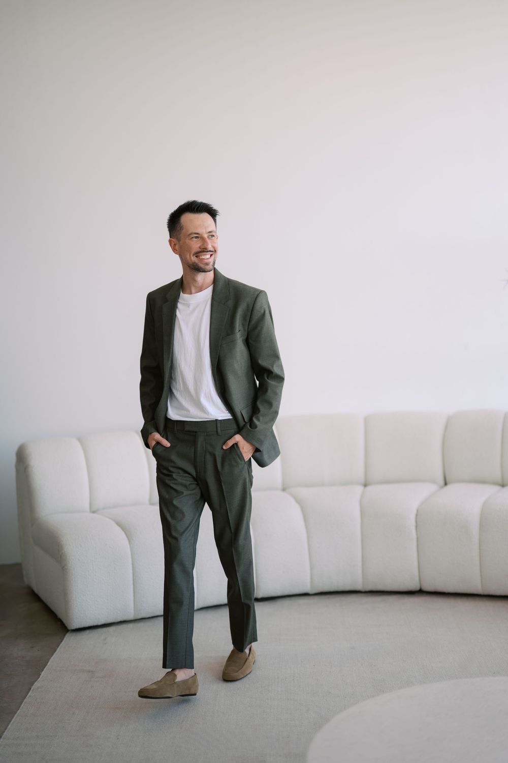 A person in a dark green suit and white t-shirt poses casually against a white sofa in a minimalist setting.