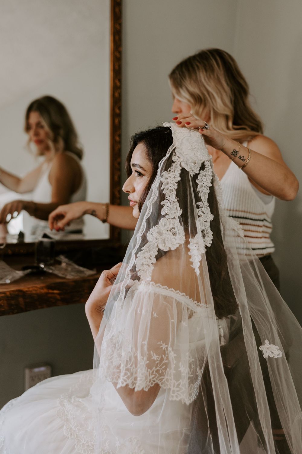 Bride in elegant lace wedding gown having her floral veil carefully placed by bridesmaid before ceremony.