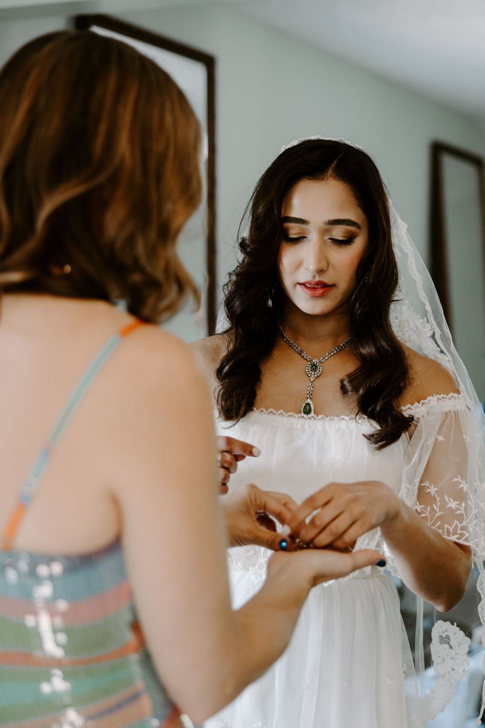 Beautiful bride in off-shoulder wedding dress with blue gemstone necklace being assisted by bridesmaid while getting ready.