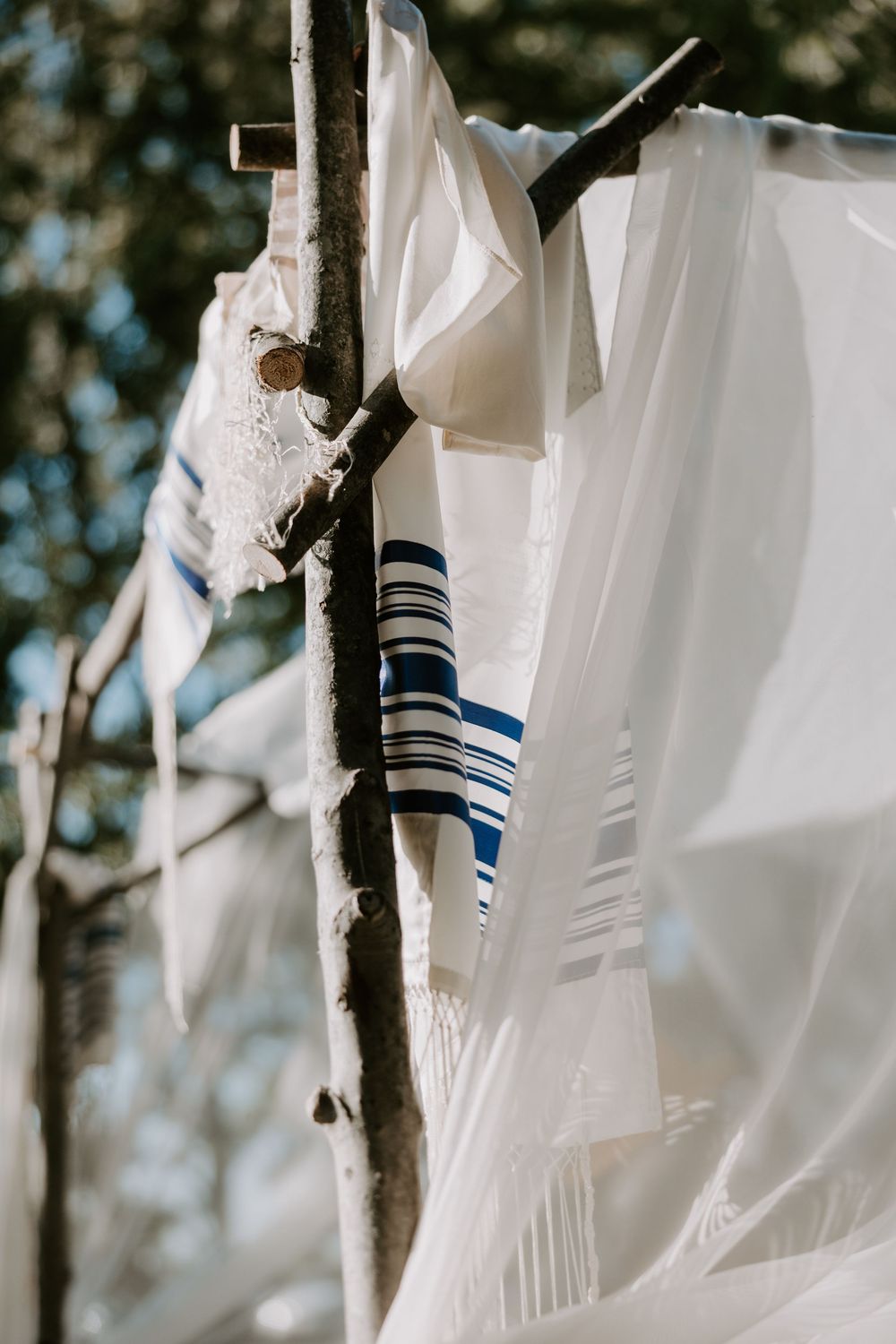 Traditional Jewish tallit prayer shawls draped over rustic wooden chuppah branches at outdoor wedding ceremony.