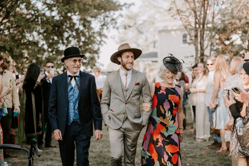 Groom in gray suit and tan hat walks down aisle with parents at outdoor bohemian wedding ceremony.