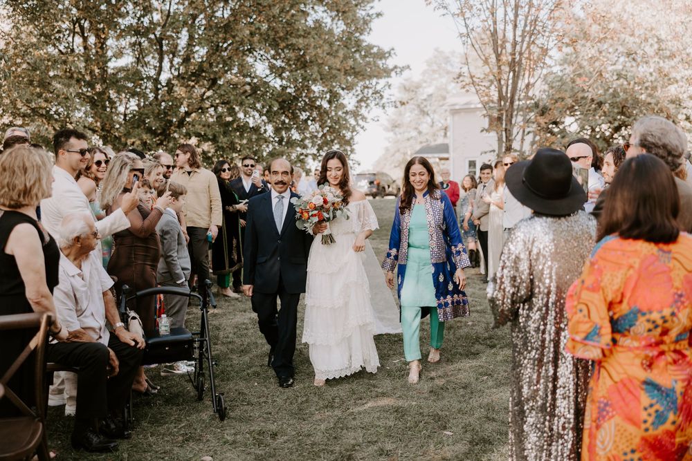 Bride in white lace gown walks down outdoor aisle with escort at rustic bohemian wedding ceremony.