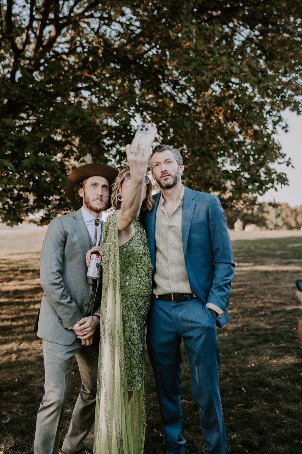 Wedding guests take selfie outdoors, man in gray suit and hat poses with woman in green dress.