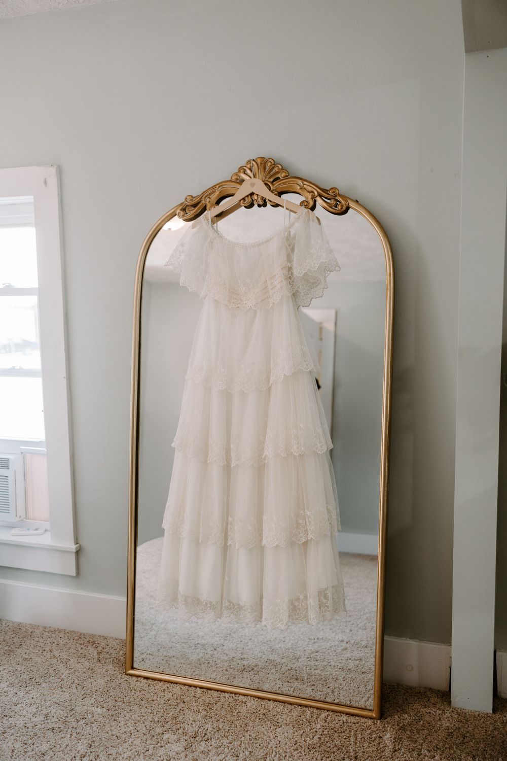 Tiered lace wedding dress displayed on a hanger reflected in an ornate gold arch mirror in a softly lit room.