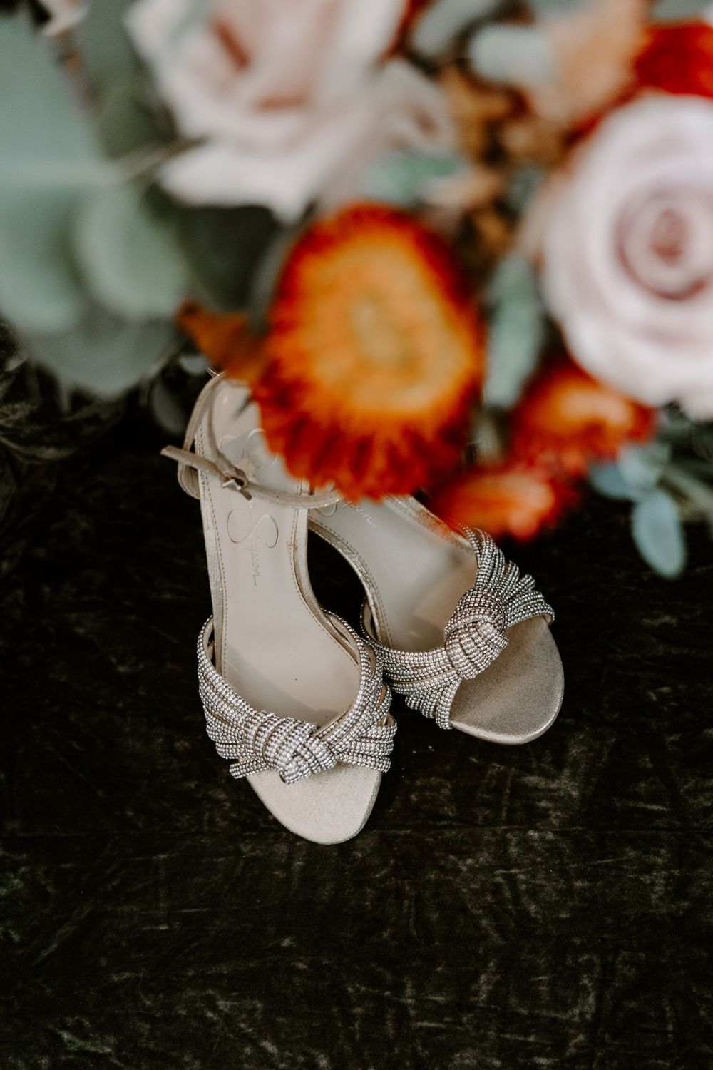 Elegant crystal-embellished bridal heels displayed in front of a colorful wedding bouquet with red and pink blooms.