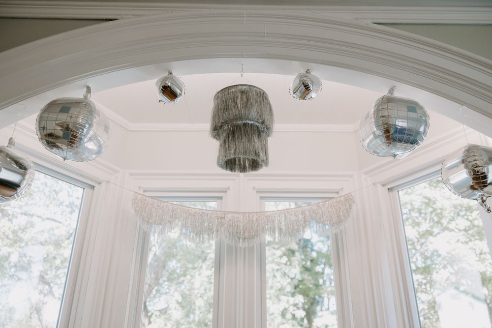 Elegant wedding reception room featuring a tiered fringe chandelier and silver disco balls hanging near arched windows.