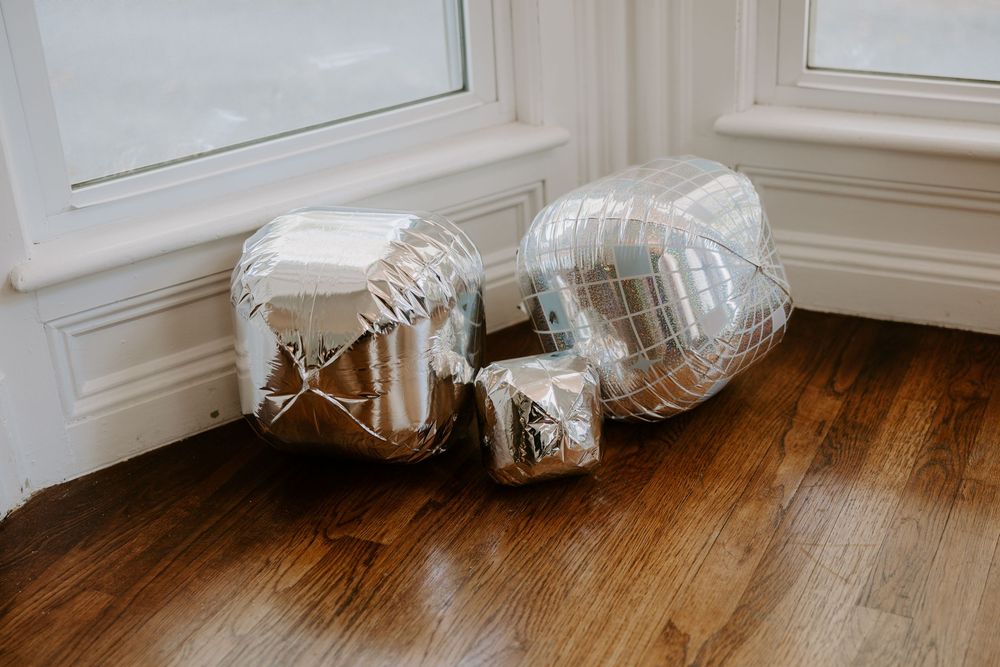 Silver metallic star and heart-shaped balloons resting on a hardwood floor near white-framed windows.