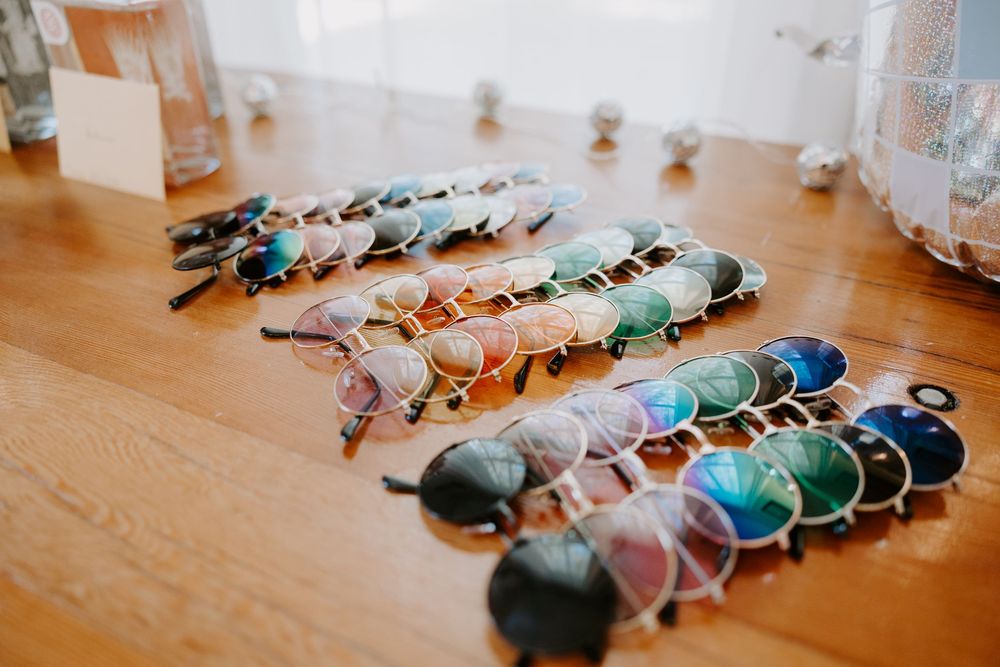 Colorful assortment of round and oval sunglasses arranged in rows on a wooden table as wedding party favors.