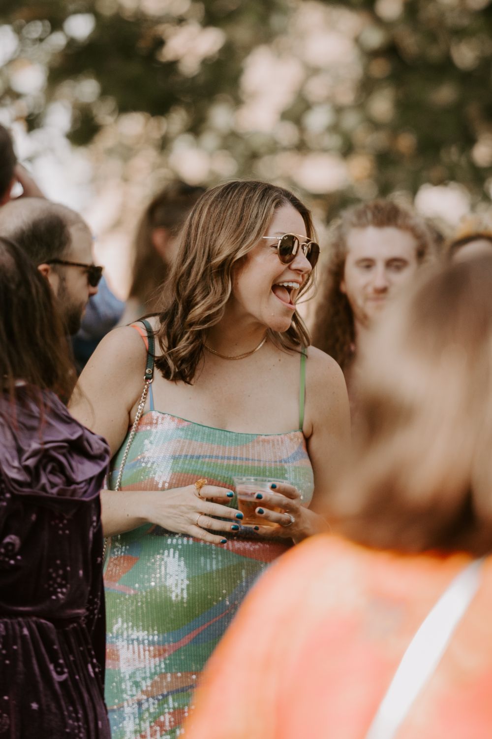 Joyful wedding guest in a green sequin dress laughing and holding a drink during an outdoor reception.