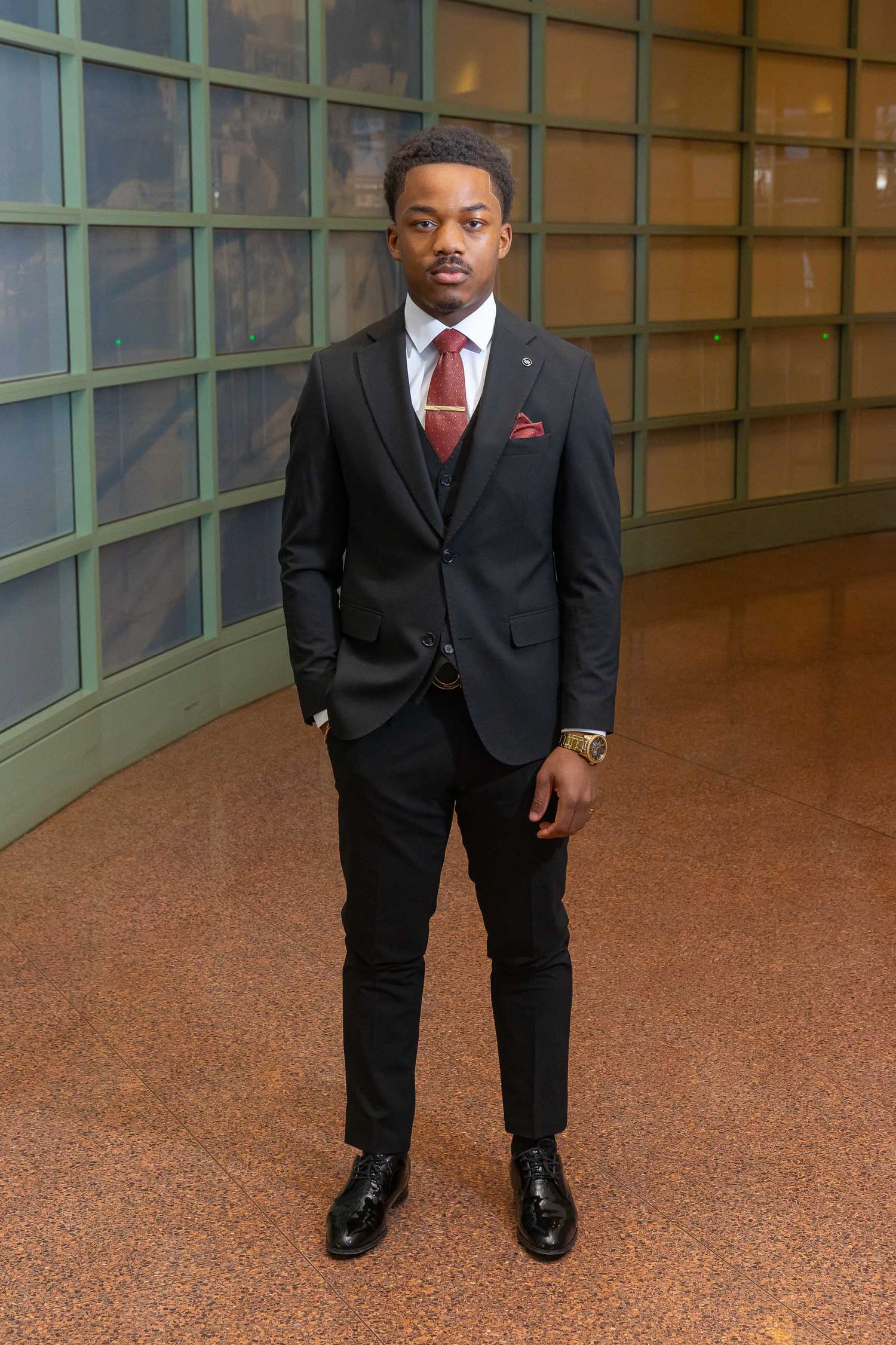 A well-dressed person in a black suit with red tie stands in front of a curved window wall in a professional setting.
