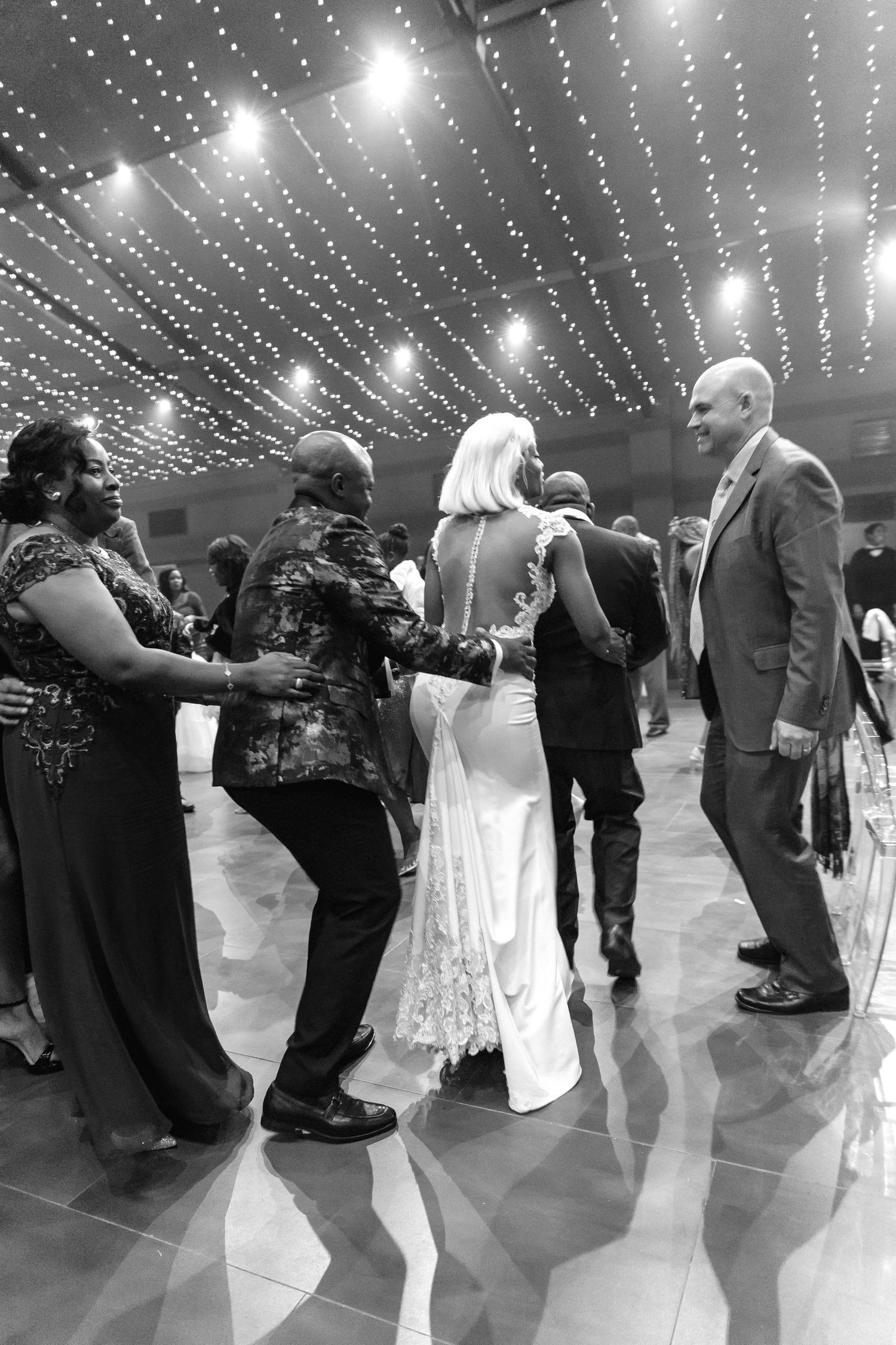 Black and white photo of elegantly dressed guests dancing and socializing at a wedding reception with twinkling lights overhead.