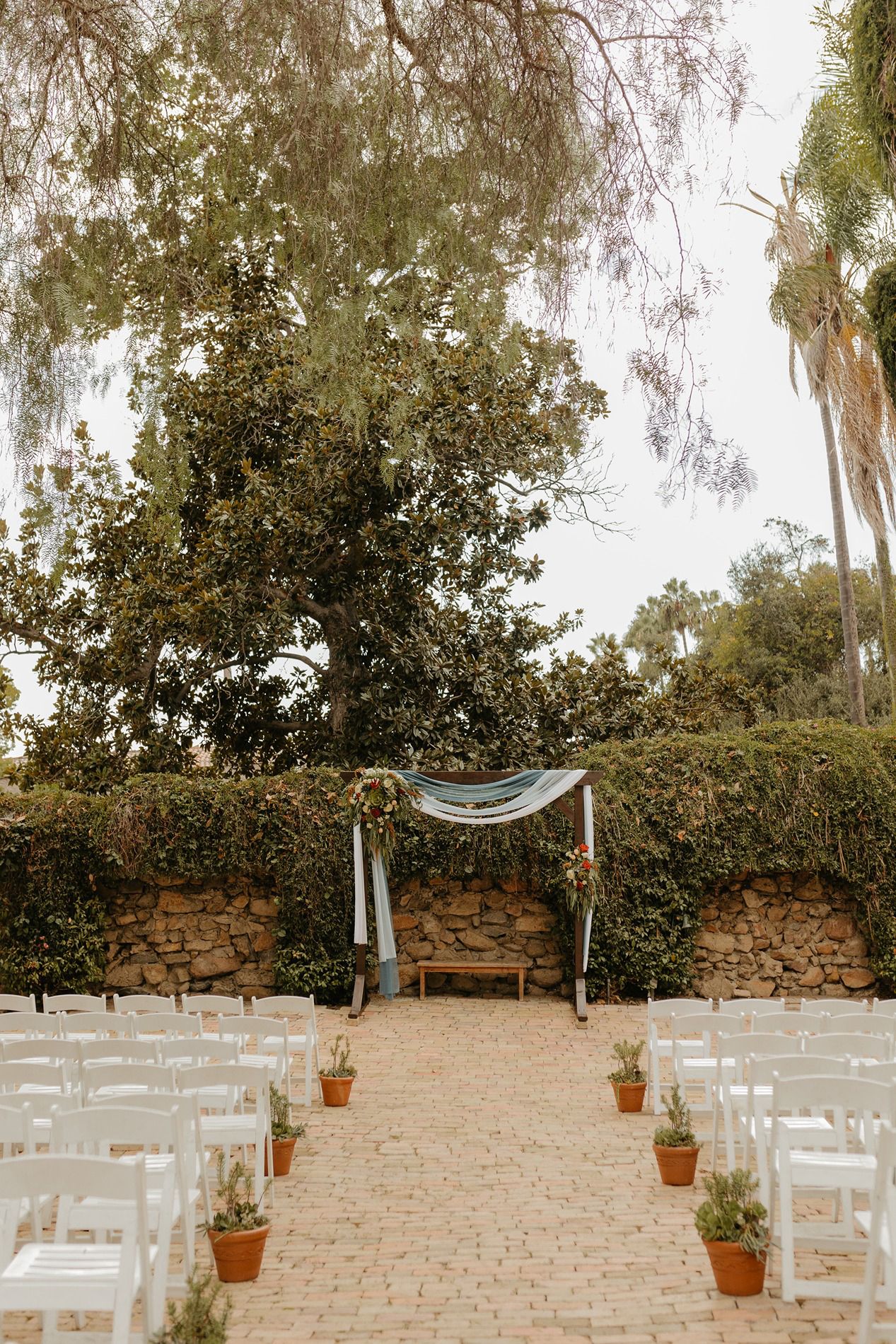 Outdoor wedding ceremony setup with white chairs, brick aisle, floral arch, and lush greenery backdrop.