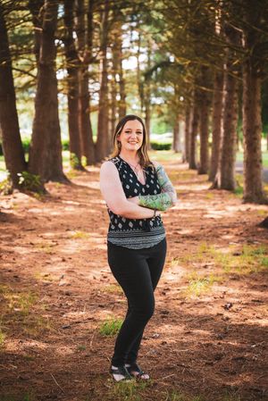 Woman with tattoos stands arms crossed among tall trees on a forest path, wearing black pants and patterned top.