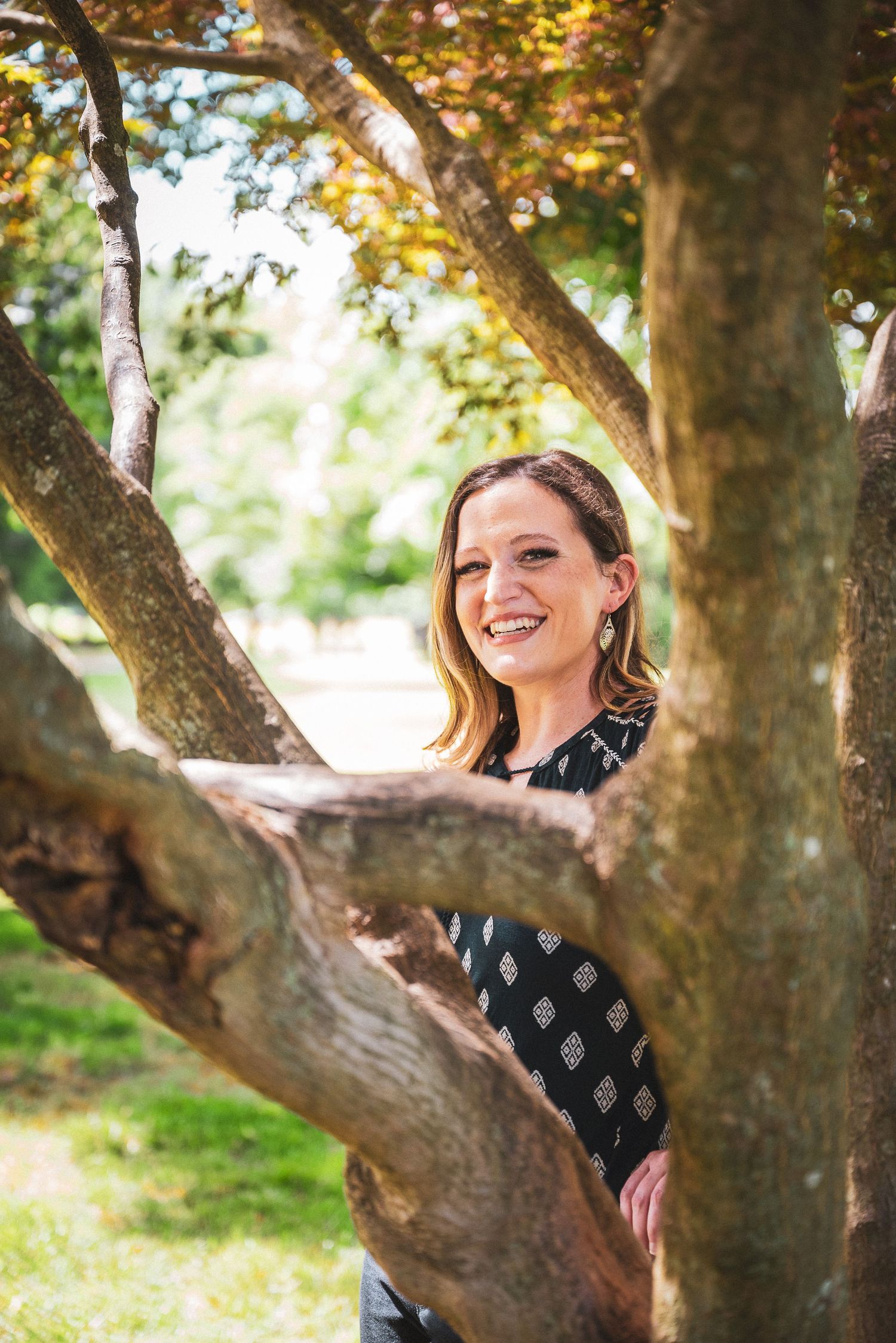 Smiling woman standing among tree branches in a sunny park, wearing a black patterned top.