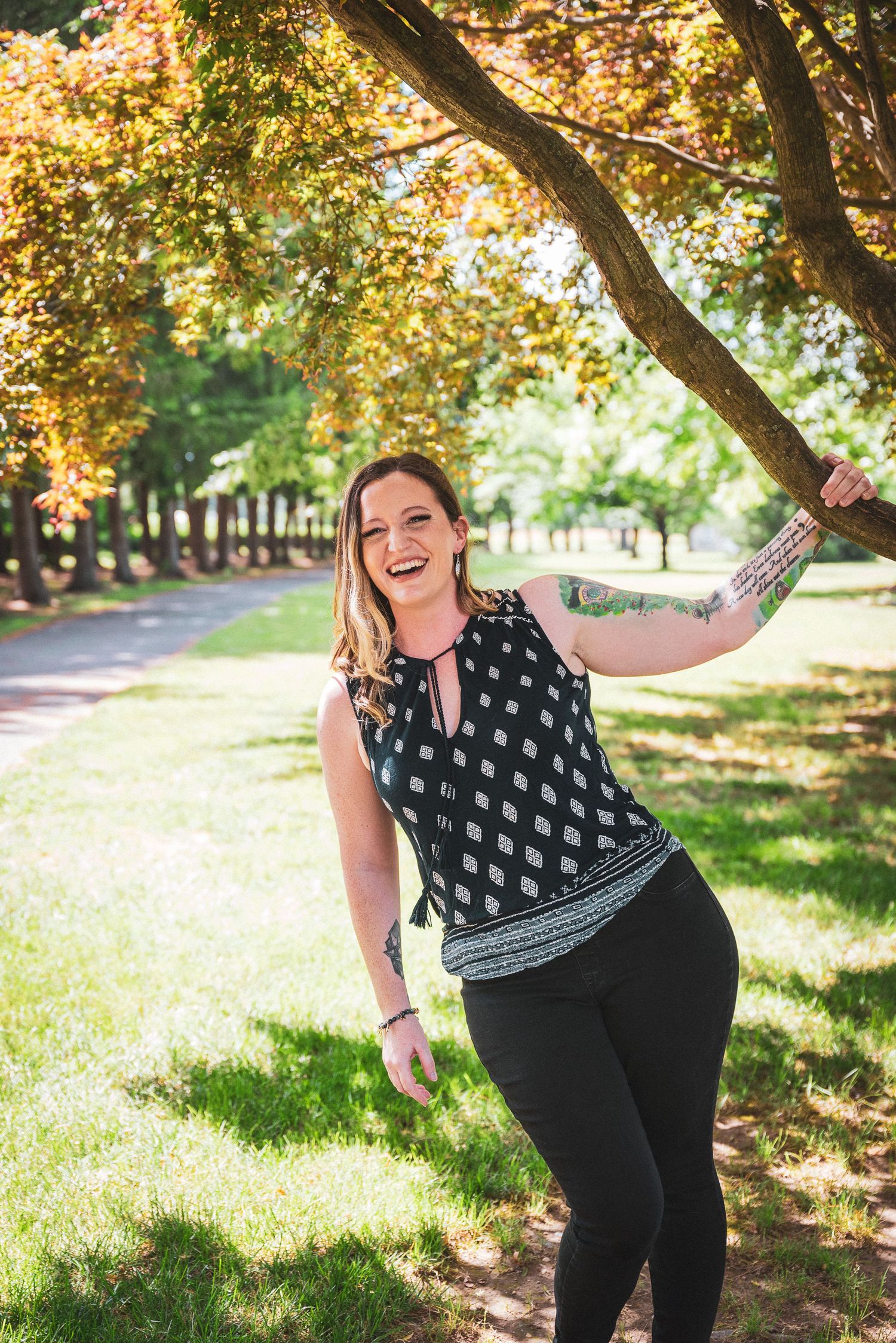 Smiling woman with tattoos holds tree branch in sunny park, wearing black patterned top and leggings.