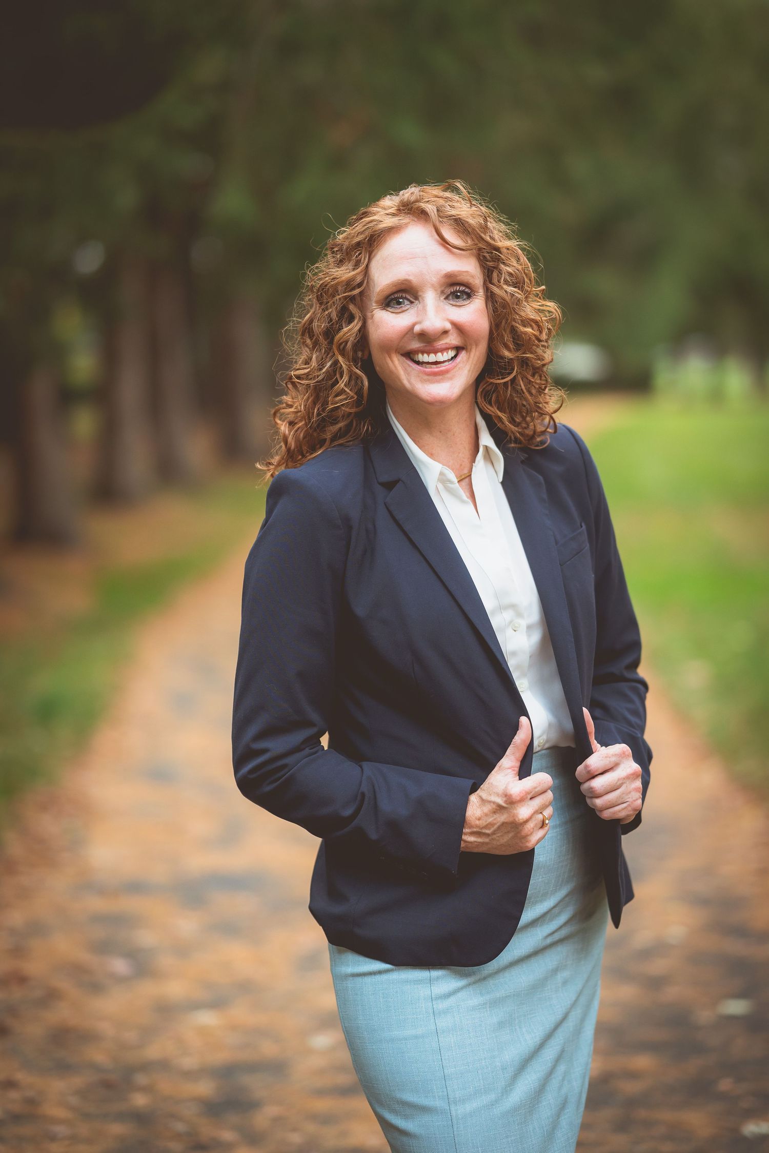 Professional woman in navy blazer and light blue skirt smiling confidently on a tree-lined path outdoors.