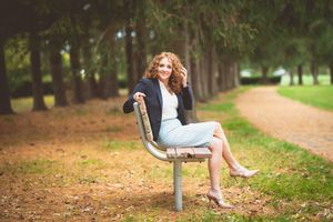 Woman with curly hair sitting on a park bench in a blue skirt and blazer, smiling outdoors.