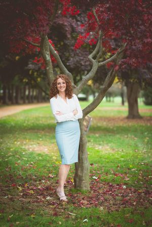 Woman in white blouse and light blue skirt standing confidently by a tree in a park with red autumn foliage.