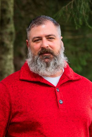 Man with gray beard in red sweater smiling outdoors among green trees.