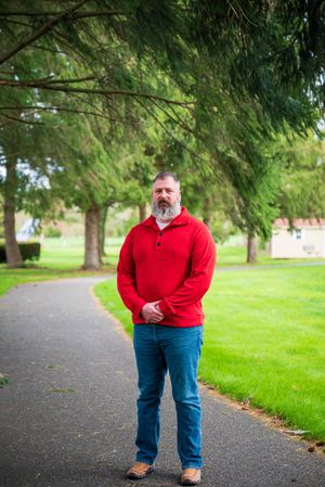 Bearded man in red sweater and jeans standing on a tree-lined path in a lush green park.