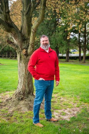 Smiling bearded man in red sweater and jeans standing by a tree in a lush green park.