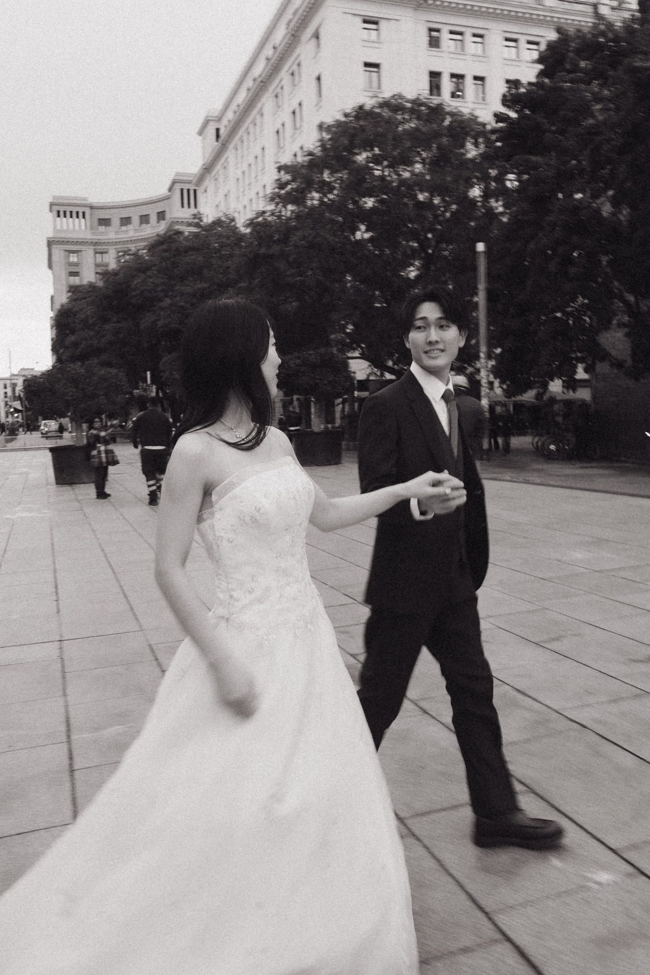 A couple in wedding attire dance together on a plaza with historic buildings in the background.