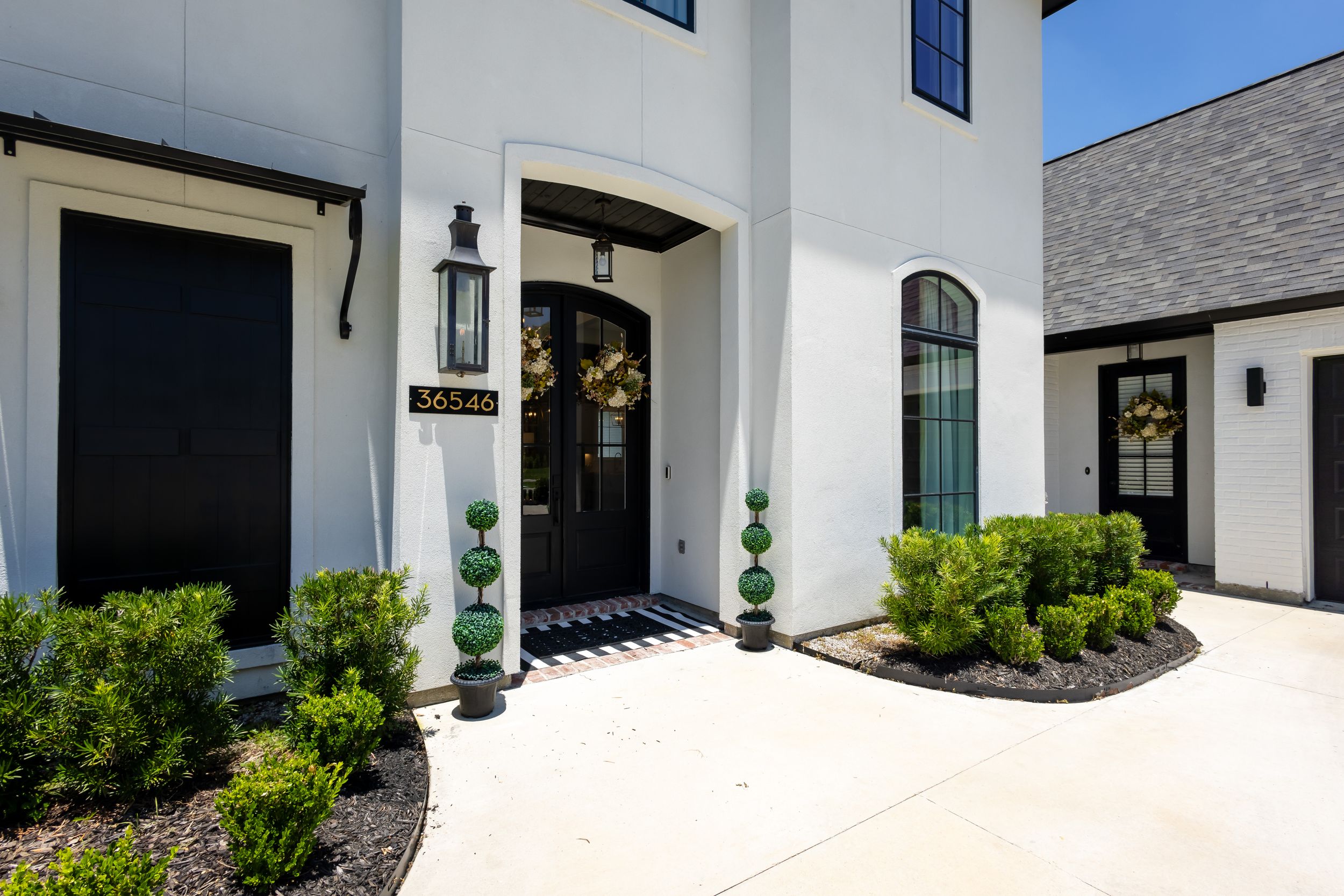 Modern white house exterior with black doors, arched entryway, and manicured landscaping featuring topiary shrubs.