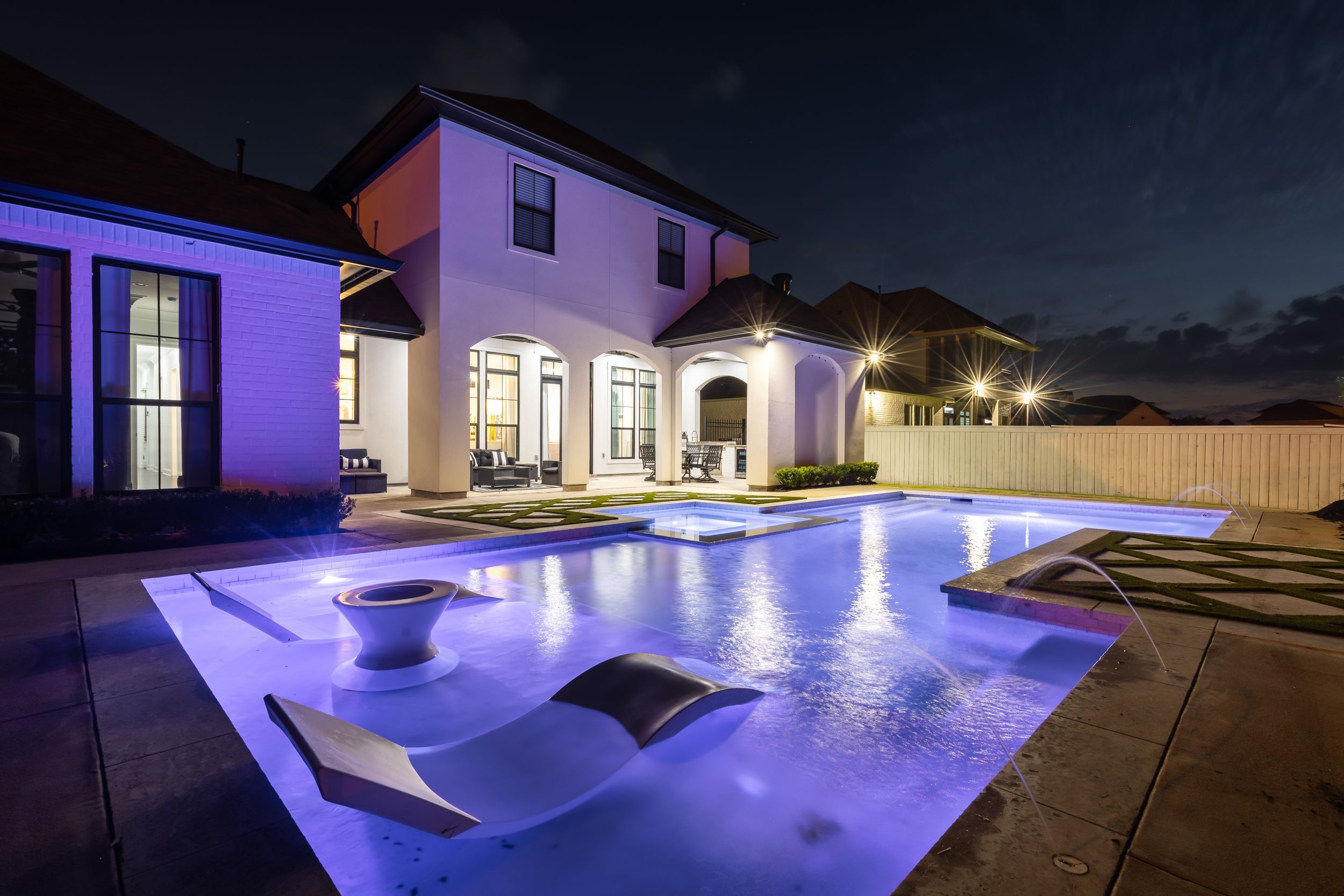 Luxurious backyard pool illuminated with purple LED lighting creates a stunning nighttime ambiance at an upscale residence.