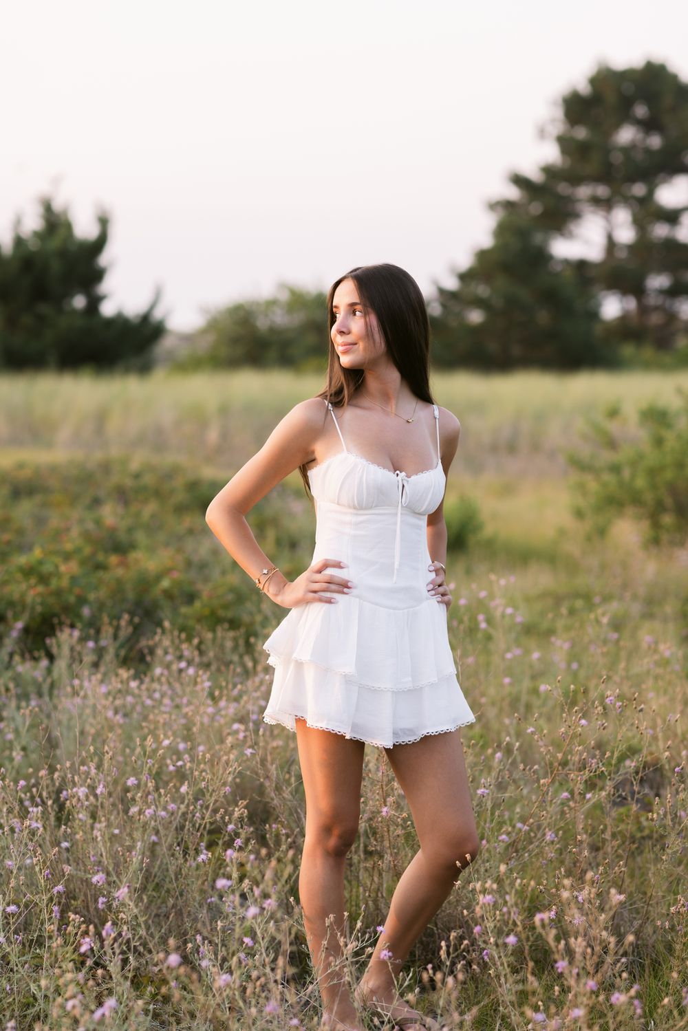 Young lady looks over shoulder toward sunset wearing a short white dress standing in purple clover for senior portraits. Newburyport senior