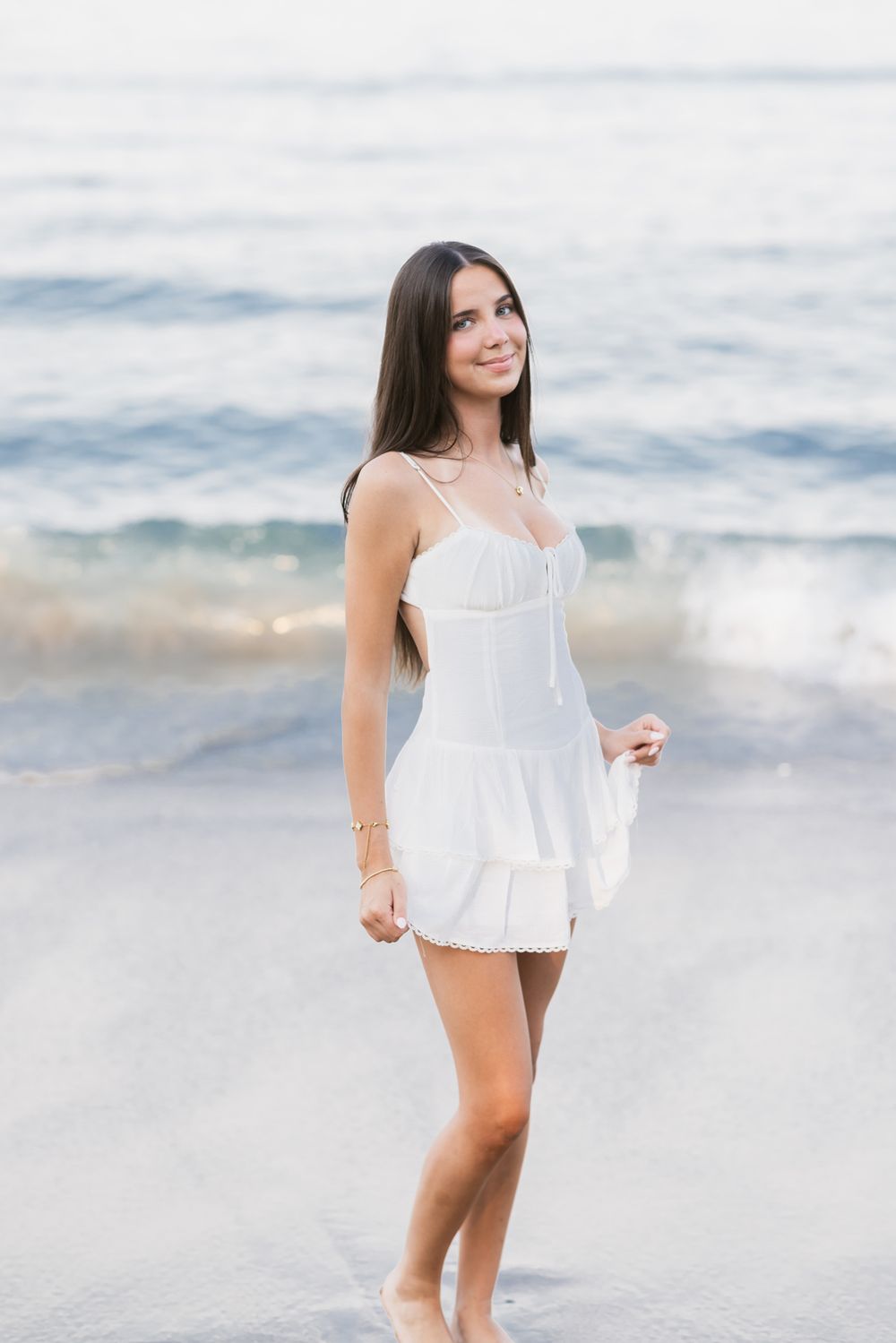Young lady with brown hair wears a short white mini dress posing in shallow ocean water at Salisbury Beach for senior photos.