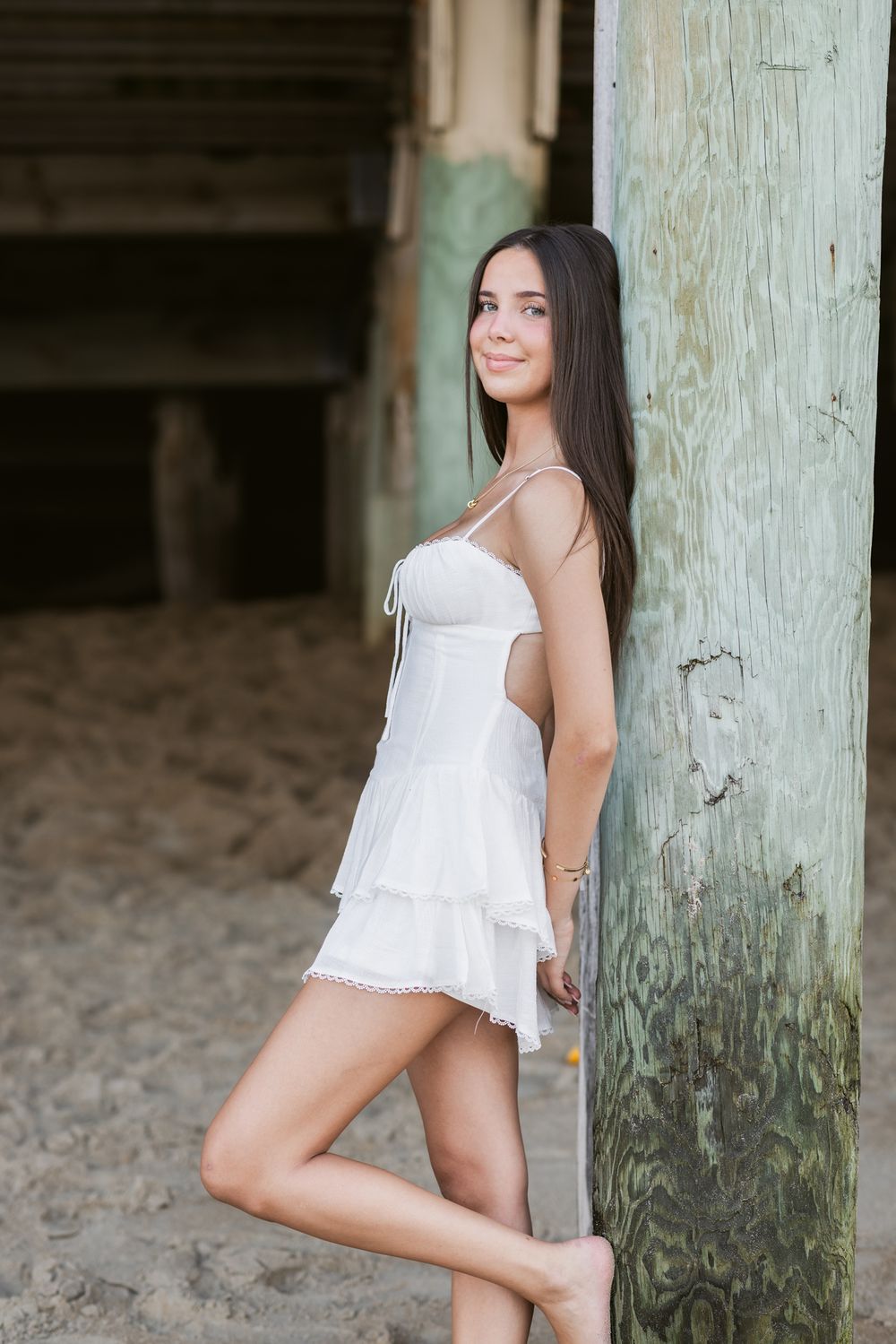 Lady wears white short dress leans on post under a boardwalk at Salisbury Beach. Seacoast New Hampshire, Southern Maine senior photographer