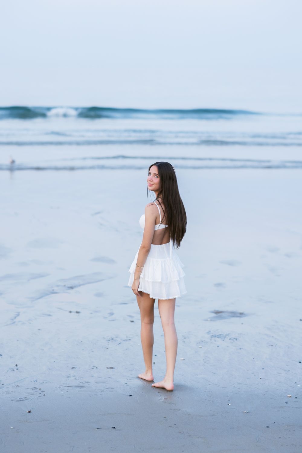 Young lady with long dark hair wearing a short white dress walks in ocean looking over shoulder at dusk. Salisbury senior photographer.