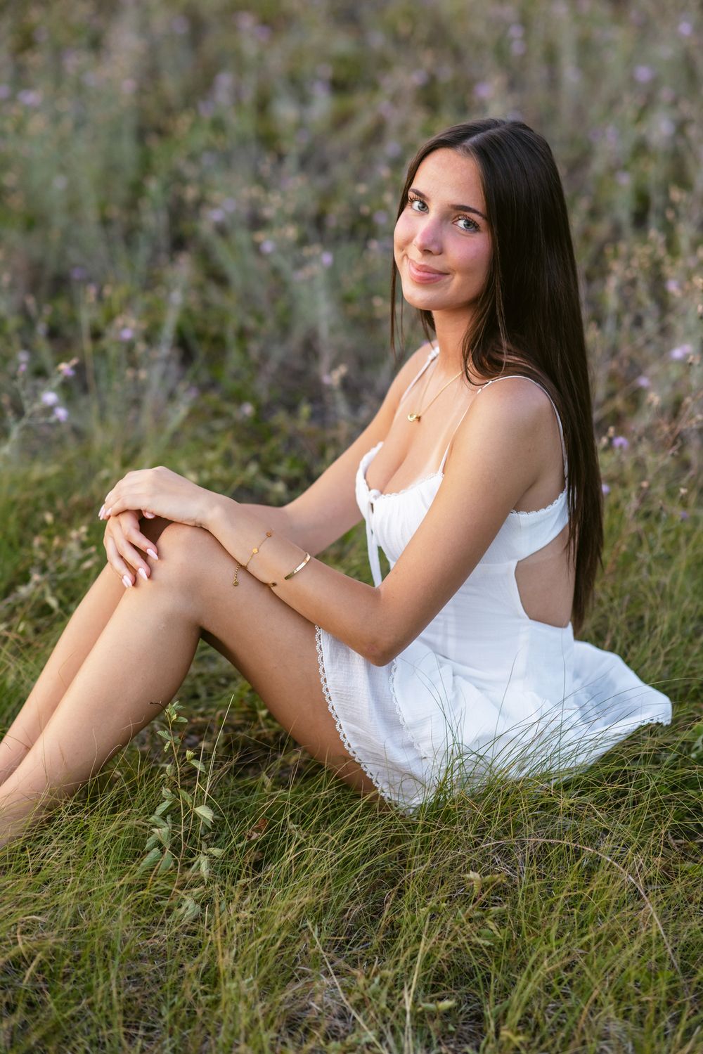 Smiling young lady in short white dress sits amid clover for senior photos. Seacoast New Hampshire, Southern Maine senior photographer.
