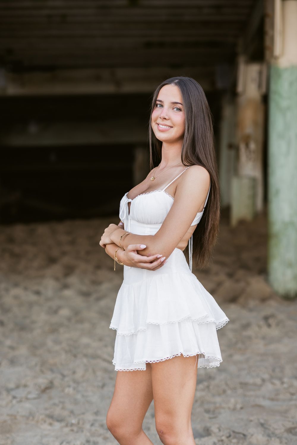 Lady wears white dress embraces arms standing under a boardwalk at Salisbury Beach for senior photos. Newburyport senior photographer.