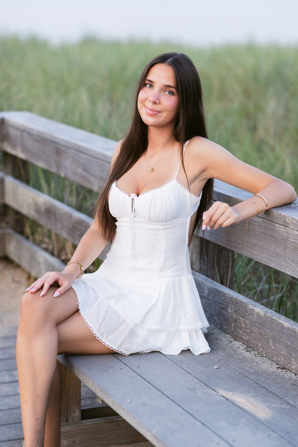 Lady wears a white short dress sits on weathered beach boardwalk for senior photos. Southern Maine New Hampshire senior photographer.