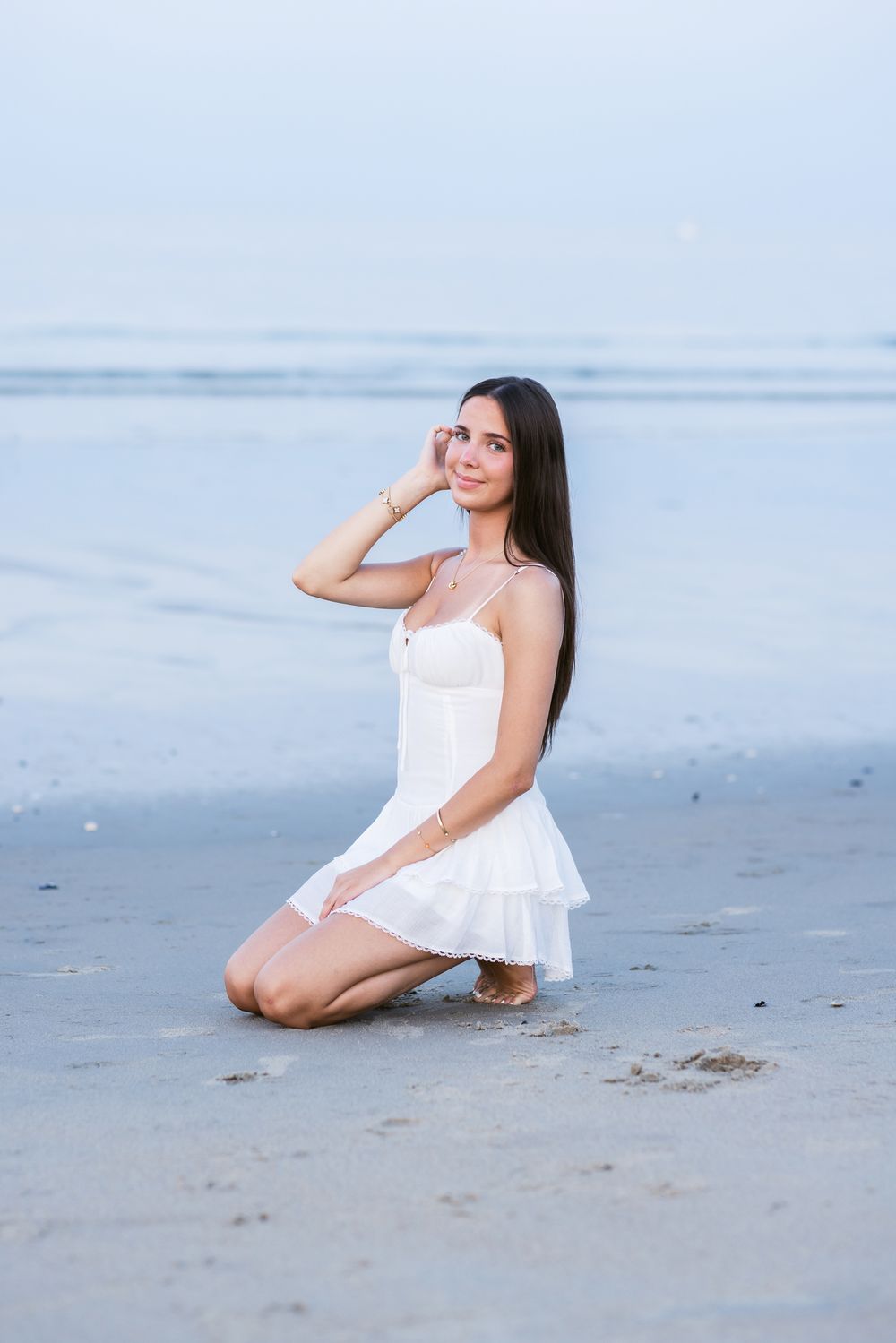 Young lady with dark hair wears a short white dress sits in sand at dusk for senior photos. Hampton, Rye, Kennebunkport senior photographer