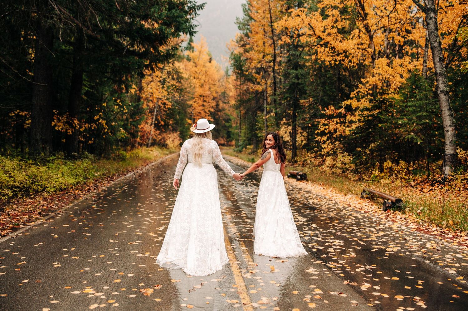 Two brides in white lace gowns hold hands on a rain-soaked road surrounded by vibrant autumn foliage.