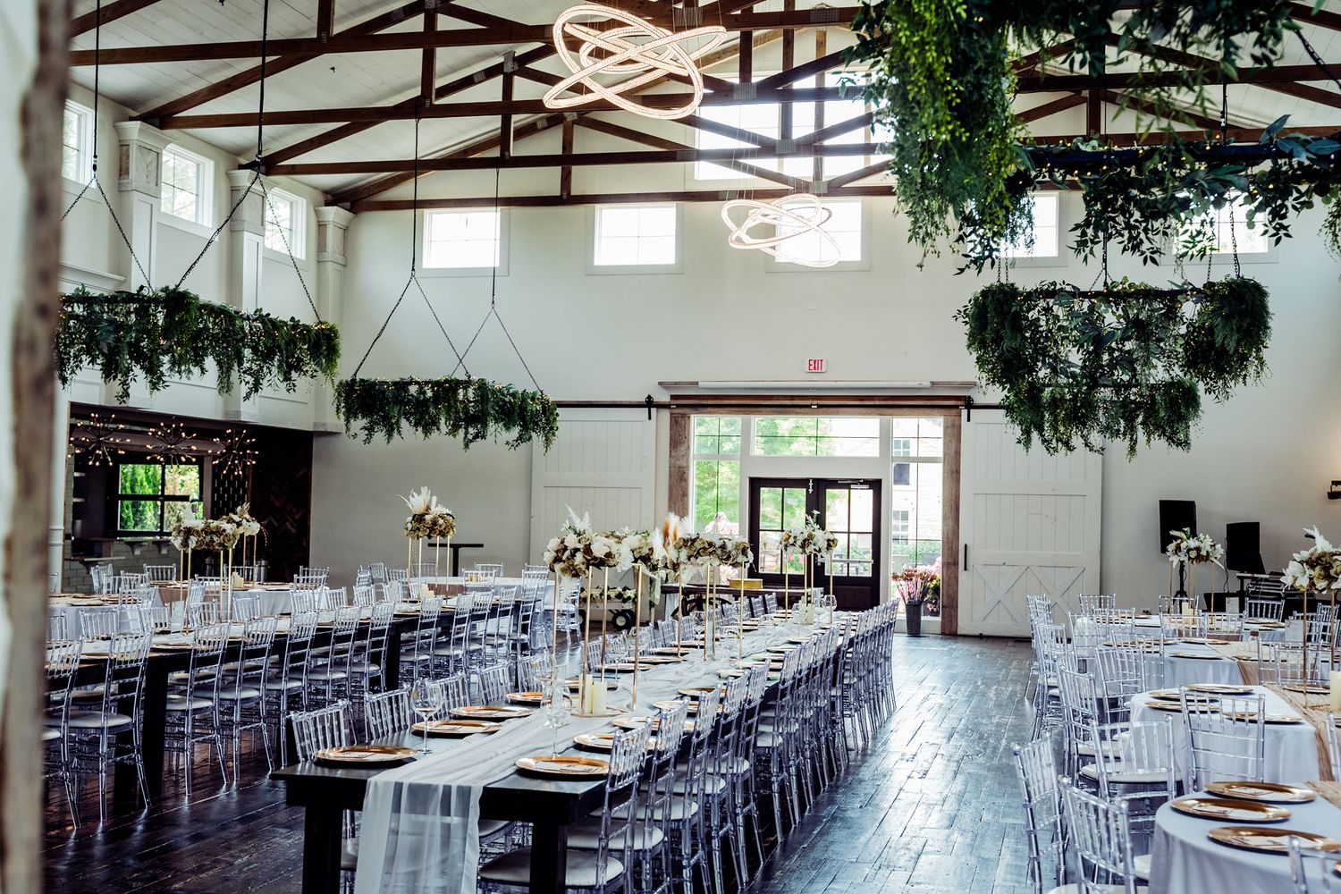 Elegant wedding reception hall with long tables, ghost chairs, hanging greenery installations, and soaring cathedral ceilings.