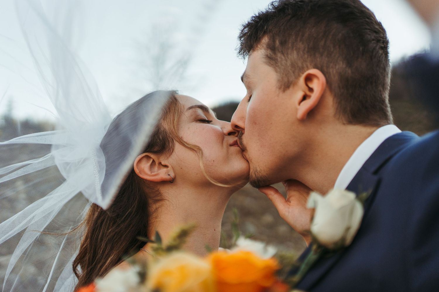 Bride and groom share a passionate first kiss under flowing veil with colorful bouquet.