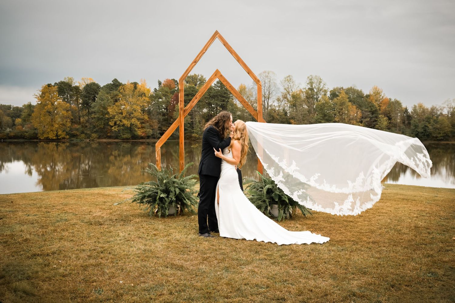 Bride and groom embrace before geometric wood arch by lake, veil flowing in wind, autumn trees backdrop.