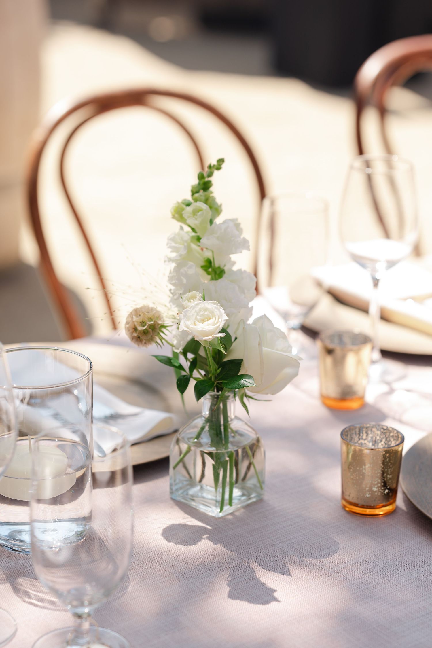 Elegant outdoor wedding reception table settings with white floral centerpieces and wooden chairs in natural lighting.