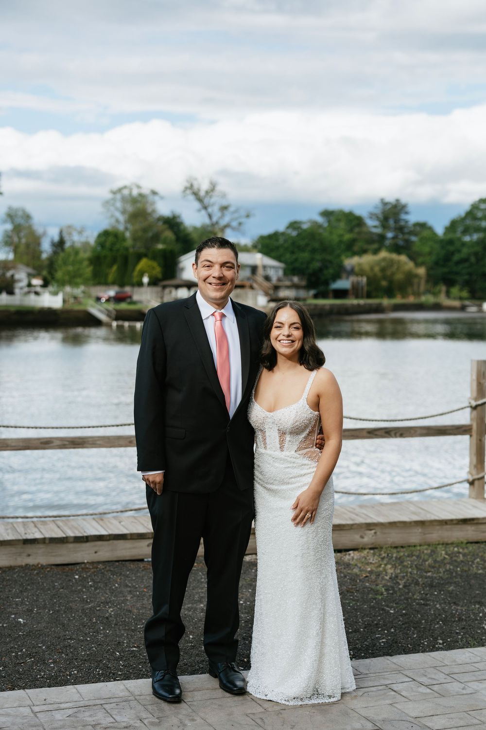 A couple poses by a lakeside on their wedding day, with water and trees visible in the background.