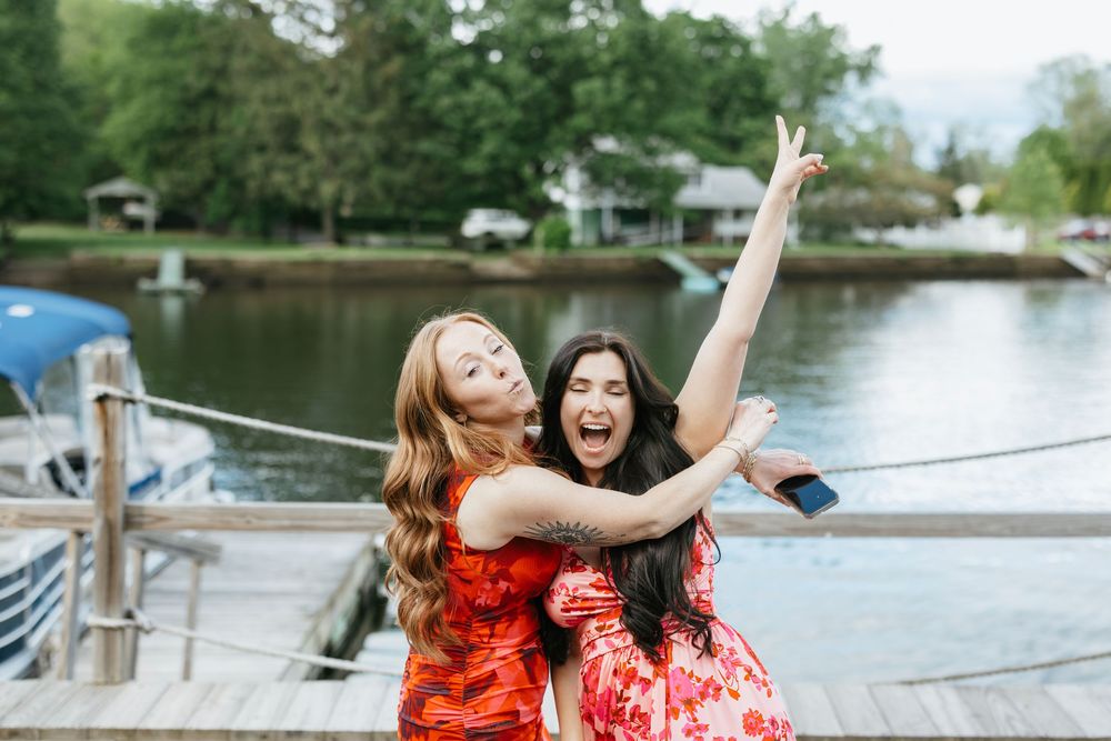 Friends in matching red floral dresses pose joyfully on a wooden dock by a serene lake.