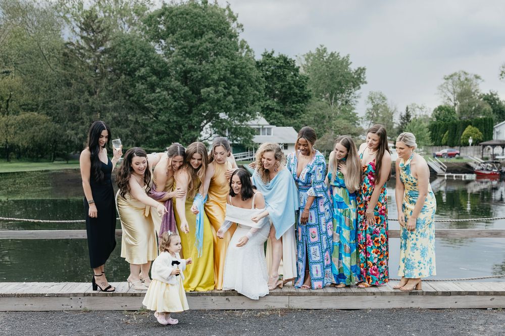 A group in colorful dresses and formal attire poses together on a dock by a peaceful lake on a summer day.