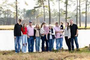 Extended family gathering posed in a line outdoors with pine trees and a lake in the background.