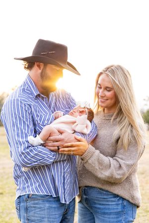 A couple in western attire shares tender moments with their newborn baby in a sunlit outdoor setting.