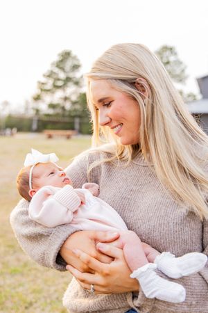 A woman in a gray sweater cradles and gazes lovingly at a newborn baby outdoors.
