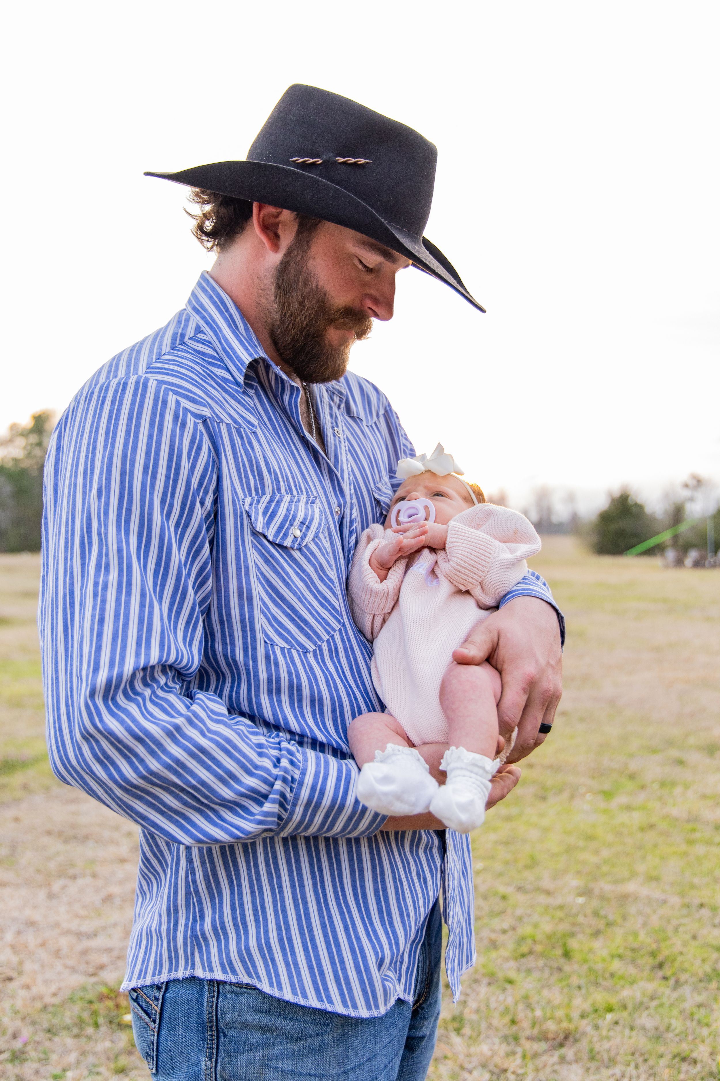 A man in a cowboy hat and striped shirt tenderly holds a small infant in an outdoor field.