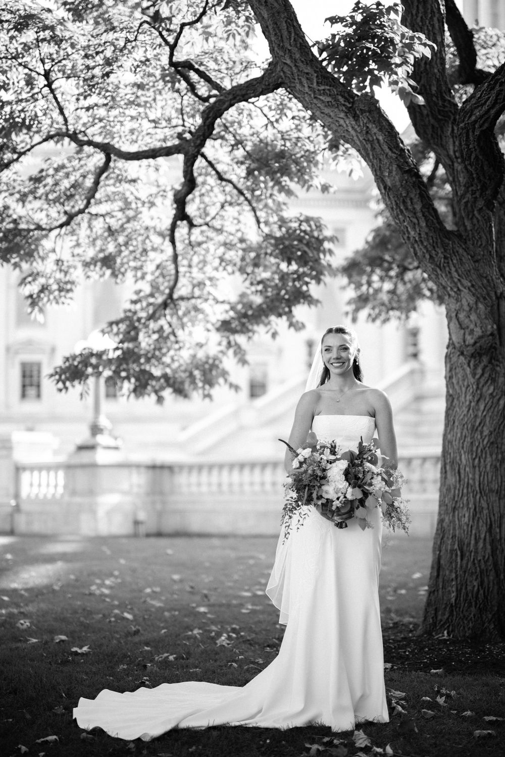 Elegant bride in white dress poses under large trees for artistic black and white wedding portraits.