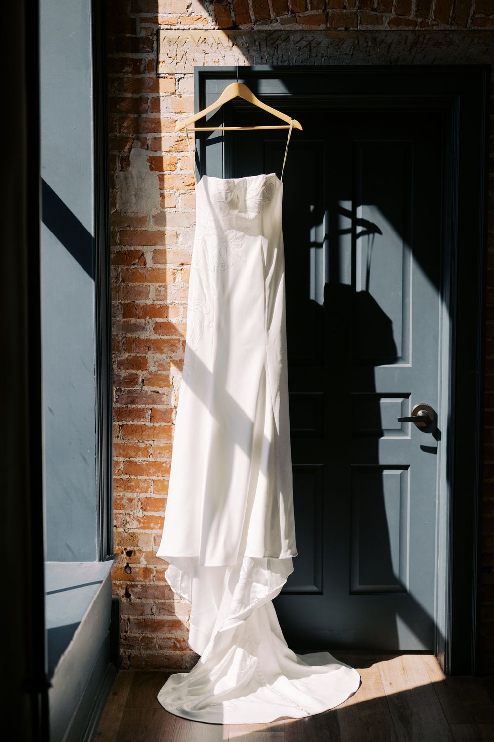 White wedding dress hanging on wooden hanger against exposed brick wall in moody lighting.